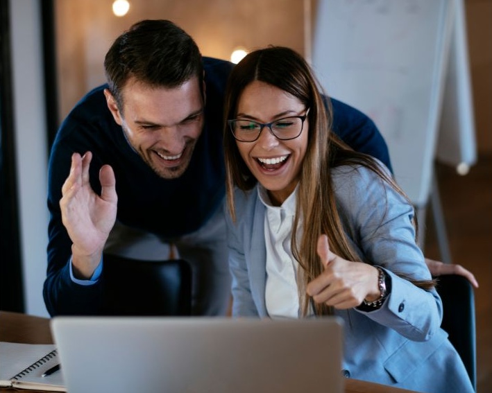 Mann und Frau vor dem Laptop und begrüßen die Menschen, die sie auf ihrem Bildschirm sehen. Beide lachen, sie hält den linken Daumen hoch. Er winkt in die Kamera.